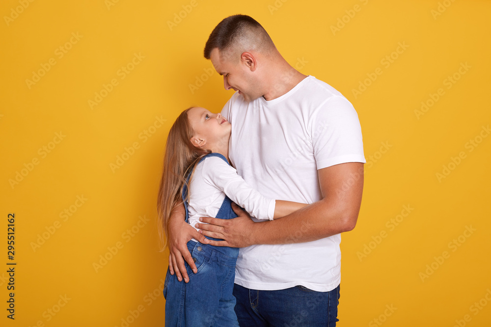 Indoor shot of young handsom man and his daughter hugging and looking at each other, stand smiling isolated over yellow studio background, wearing casual clothing. Family and relationship concept. Indoor shot of young handsom man and his daughter hugging and looking at each other, stand smiling isolated over yellow studio background, wearing casual clothing. Family and relationship concept.