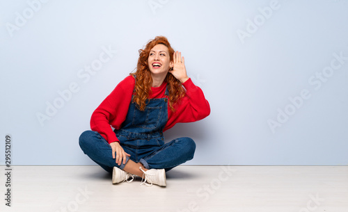 Redhead woman with overalls sitting on the floor listening to something by putting hand on the ear