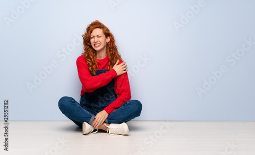 Redhead woman with overalls sitting on the floor suffering from pain in shoulder for having made an effort