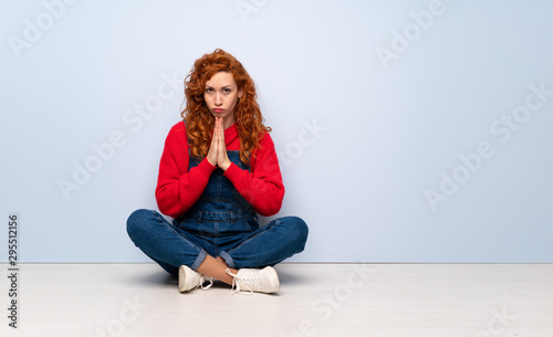 Redhead woman with overalls sitting on the floor keeps palm together. Person asks for something