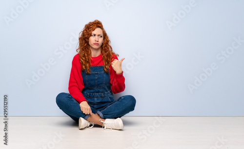 Redhead woman with overalls sitting on the floor unhappy and pointing to the side