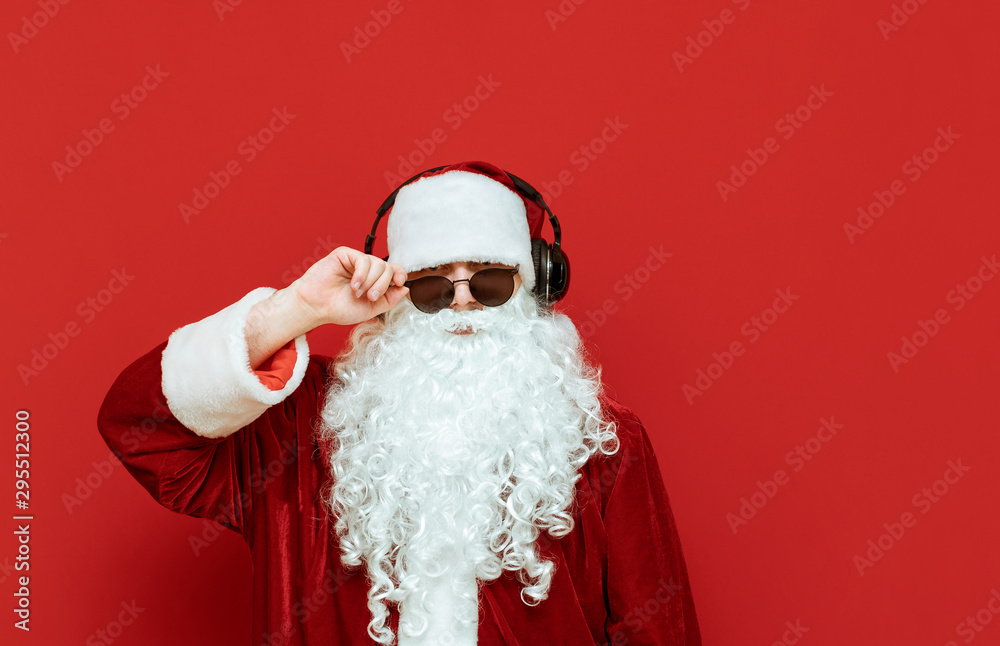 Portrait of cool Santa Claus in sunglasses and headphones posing at camera isolated on red background. Swag Santa stands on a magical background and listens to music in headphones. Christmas concept. Portrait of cool Santa Claus in sunglasses and headphones posing at camera isolated on red background. Swag Santa stands on a magical background and listens to music in headphones. Christmas concept.