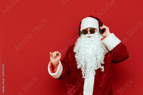 Studio photo of cool Santa Claus in sunglasses having fun on red background, listening to music in headphones and dancing. Santa at New Year's party. Copyspace photo