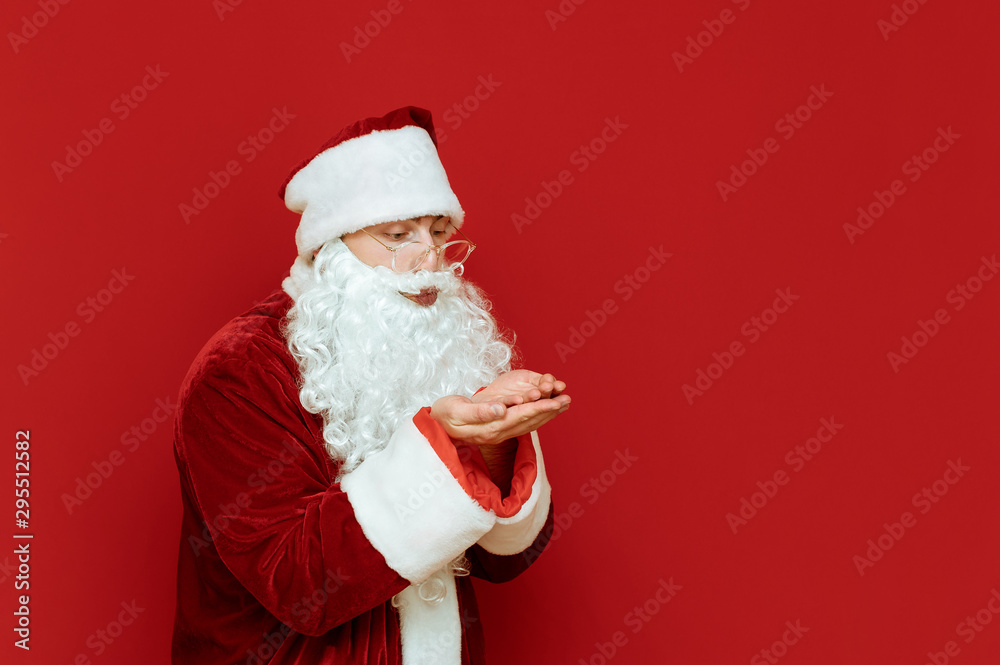 Portrait of Santa standing on red background looks at raised hands and sends an air kiss, looks away. Christmas concept. Happy xmas. Copyspace Portrait of Santa standing on red background looks at raised hands and sends an air kiss, looks away. Christmas concept. Happy xmas. Copyspace