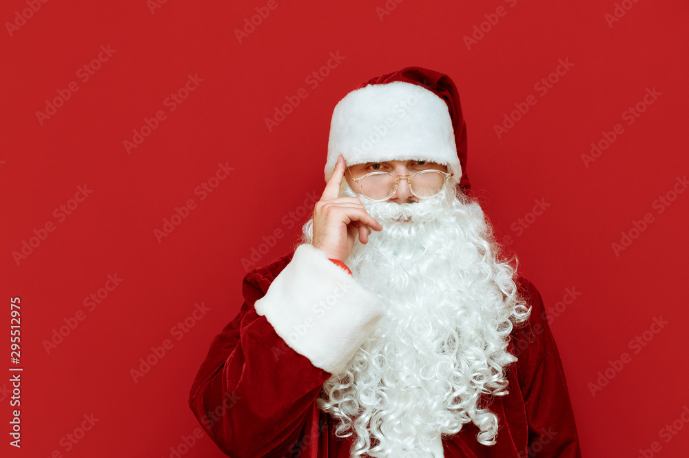 Portrait of Santa Claus shows gesture think about it, holds his finger near his head and looks into the camera with a serious face. Pensive santa isolated on red background. Christmas concept. Xmas