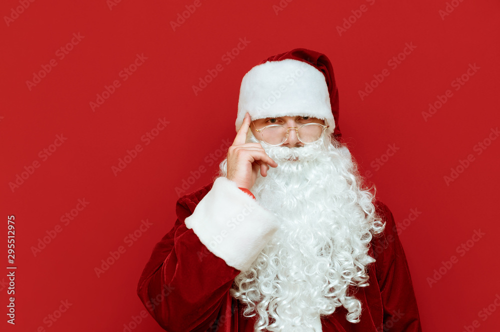 Portrait of Santa Claus shows gesture think about it, holds his finger near his head and looks into the camera with a serious face. Pensive santa isolated on red background. Christmas concept. Xmas Portrait of Santa Claus shows gesture think about it, holds his finger near his head and looks into the camera with a serious face. Pensive santa isolated on red background. Christmas concept. Xmas