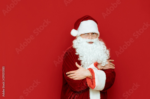 Santa Claus freezes, warms up against a red background and looks in camera with a sad face.Portrait Santa Claus who is cold. Man in santa costume freezing on red background. Cold winter for Christmas