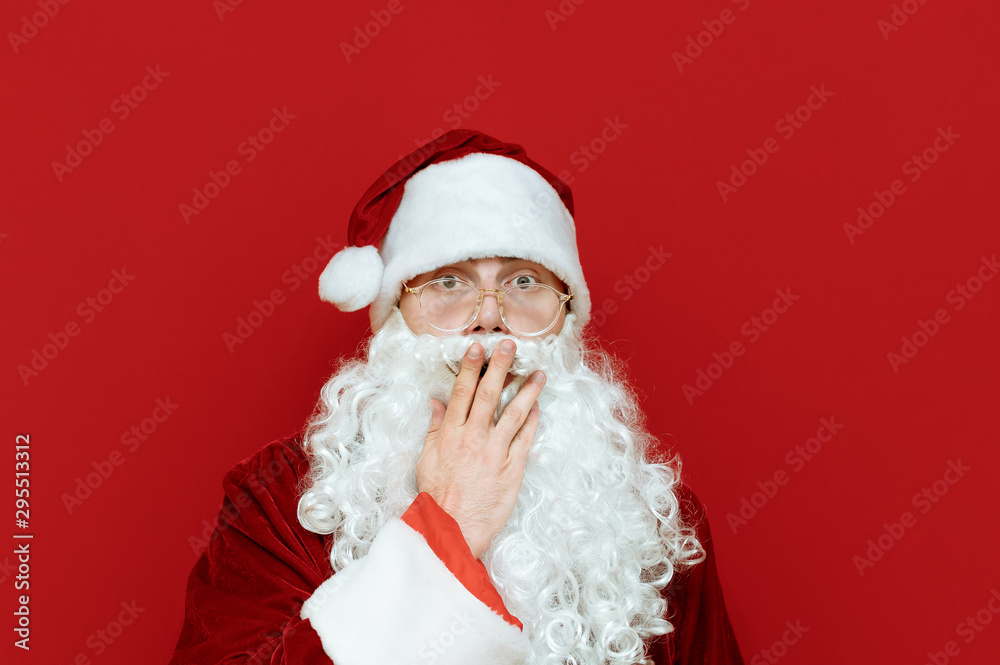 Closeup studio portrait of shocked Santa Claus on red background, covered his mouth in astonishment, looks into camera with astonished face. Emotional Santa isolated on red background Closeup studio portrait of shocked Santa Claus on red background, covered his mouth in astonishment, looks into camera with astonished face. Emotional Santa isolated on red background