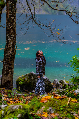 a young, red-haired girl with glasses, fashionable, sportily dressed stands in the rain, in the background an alpine lake photo