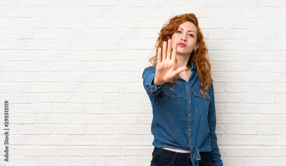 Redhead woman over white brick wall making stop gesture
