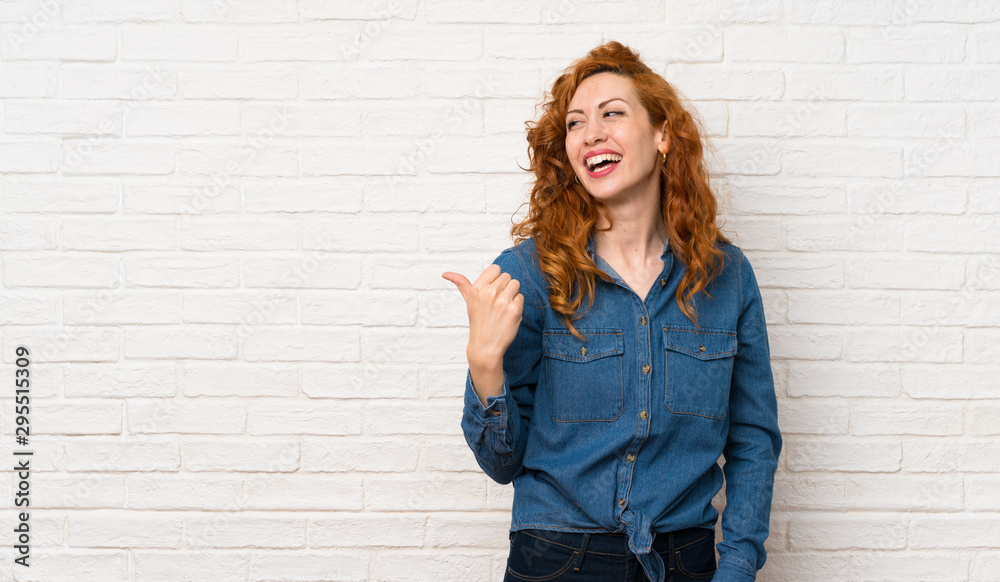 Redhead woman over white brick wall pointing to the side to present a product Redhead woman over white brick wall pointing to the side to present a product