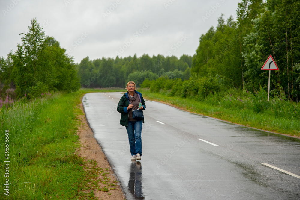 An aged woman walks along a wet highway that is in the woods.
