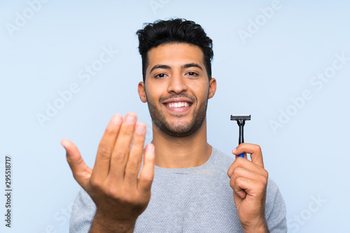 Young man shaving his beard over isolated blue background inviting to come with hand. Happy that you came