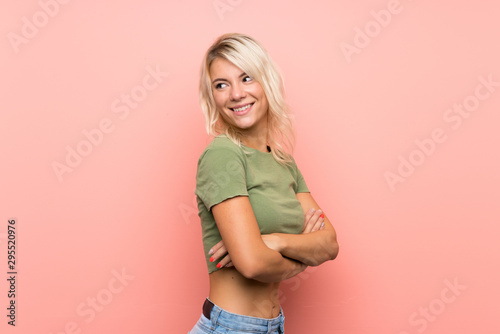 Young blonde woman over isolated pink background with arms crossed and happy