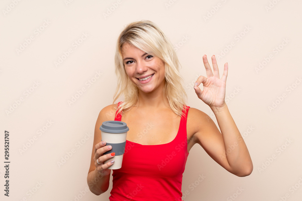 Young blonde woman over isolated background holding coffee to take away while making OK sign Young blonde woman over isolated background holding coffee to take away while making OK sign