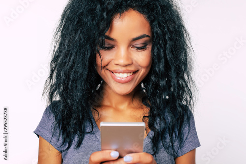 Can’t be better. Close-up portrait of Afro woman with broad alluring smile and long frizzy hair, who expresses happiness while reading something on her smartphone.