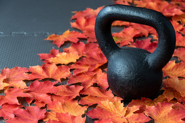 Black iron kettlebell on a black rubber gym floor, with orange and yellow maple leaves, fall fitness