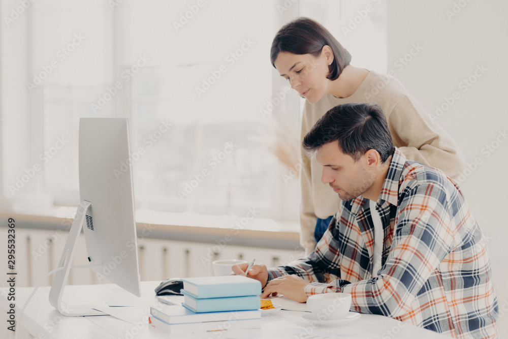 Husband and wife work on paper report together, check information, sit in front of computer, drinks coffee, dressed in casual wear, collaborate for teamworking. Family and remote work concept Husband and wife work on paper report together, check information, sit in front of computer, drinks coffee, dressed in casual wear, collaborate for teamworking. Family and remote work concept
