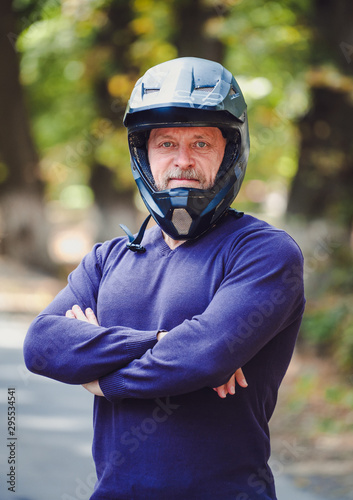 a senior man in dark helmet outdoor. Casual clothes. Crossed hands. Blue sweater. Outdoor blurred background. Closeup. © Vadim