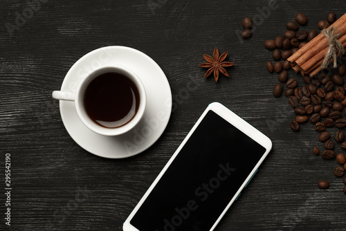 White cup with cinnamon and anise coffee sticks and a mobile phone with a blank screen on a black background with coffee beans and place for text top view