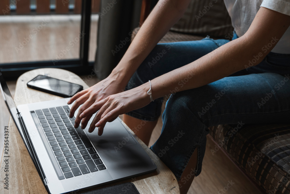 Naklejka premium Woman is sitting on a sofa and using a laptop at wooden table. Study and work online, freelance. Self employed girl is working with her notebook sitting on a couch with a phone and ereader on table.