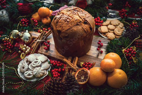 Christmas ingredients table with a Panettone in the center on a wooden board, accompanied by orange, cookies, cinnamon, pine. Festive pine decoration on red rustic background