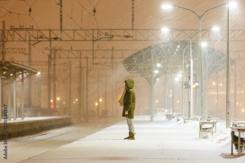 Obraz premium Young woman in hood on empty railway platform in blizzard waiting for a train. Female is waiting for the train in bad weather in the evening, strong wind. Transport delay.