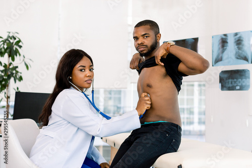 Young pretty African woman doctor examining African man patient with stethoscope in medical office