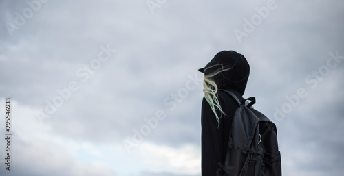 Back view of traveler on background of sky, wearing black coat and backpack.