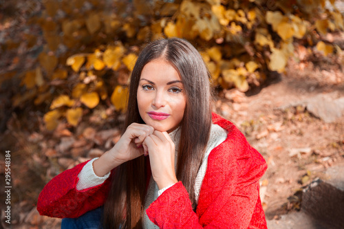 Young beautiful brunette woman with red lips and long straight hair wearing red jacket sitting outdoor in autumn park.