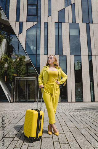 Young overjoyed traveler tourist woman in yellow summer yellow casual clothes. Girl traveling abroad on weekend getaway. Woman is dressed in yellow shoes with a yellow suitcase and black glasses.