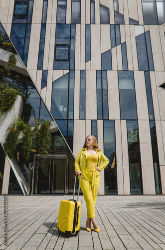 Young overjoyed traveler tourist woman in yellow summer yellow casual clothes. Girl traveling abroad on weekend getaway. Woman is dressed in yellow shoes with a yellow suitcase and black glasses.
