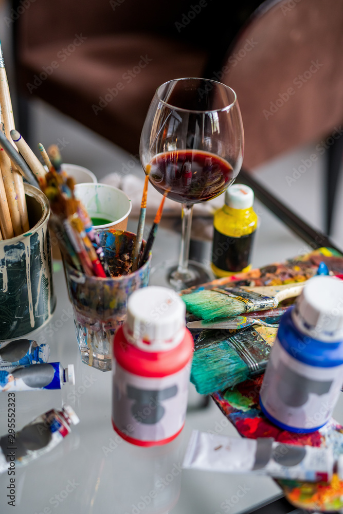 Glass of red wine among paintbrushes and various paints and gouaches