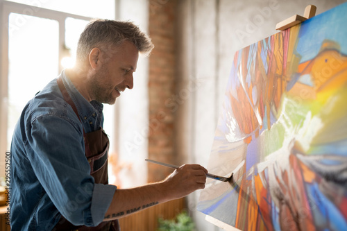 Happy mature master of arts in apron working over abstract painting photo