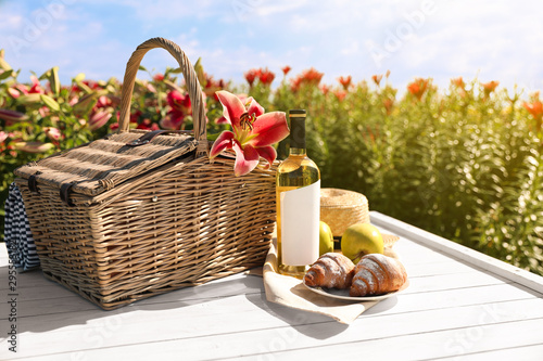 Composition with picnic basket and bottle of wine on white wooden table in lily field. Space for text