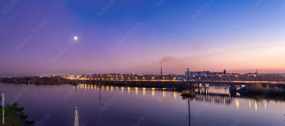 Panorama of the summer night city with a view of the railway bridge by the river. There is a moon in the sky. Lights are reflected in the water.