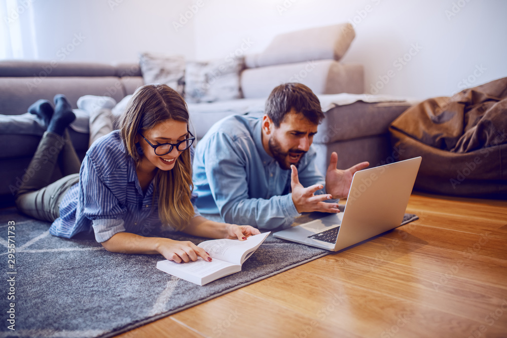 Cheerful caucasian woman lying on the floor and reading book. Her nervous boyfriend lying next to her and looking at laptop. Living room interior. Cheerful caucasian woman lying on the floor and reading book. Her nervous boyfriend lying next to her and looking at laptop. Living room interior.