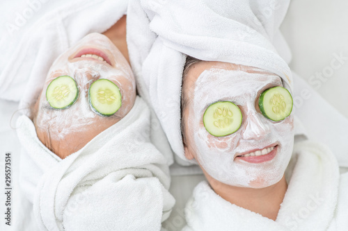 Mother and daughter in white bathrobes and with towels on their heads applying pieces of cucumber to their eyes