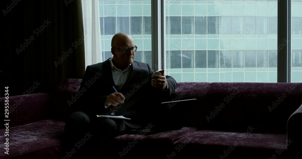 A rich man in a suit and glasses sits on the couch of his office with a laptop documents and phone and works A rich man in a suit and glasses sits on the couch of his office with a laptop documents and phone and works