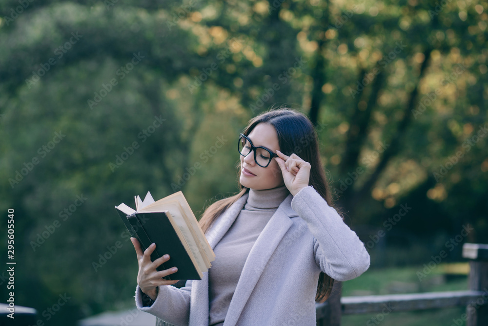 Fototapeta premium Smart brunette woman in eyeglasses reading book in park 