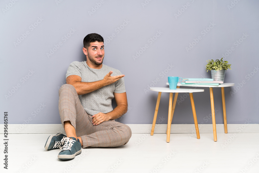 Young handsome man sitting on the floor extending hands to the side for inviting to come