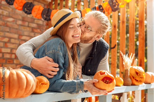 Halloween Preparaton Concept. Young couple leaning on fence decorating house with jack-o'-lantern husband hugging and kissing wife holding carved pumpkin cheerful