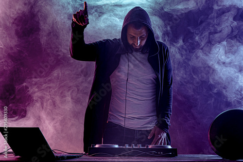 Young stylish man in glasses posing behind mixing console on colored smoke studio background.