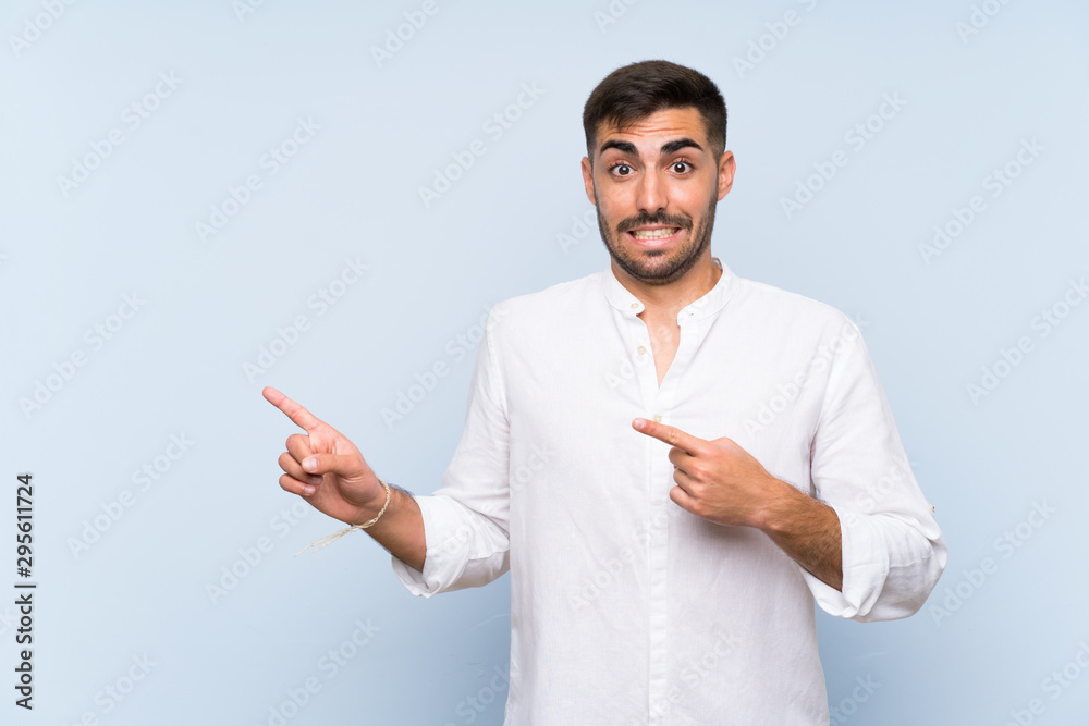 Handsome man with beard over isolated blue background frightened and pointing to the side Handsome man with beard over isolated blue background frightened and pointing to the side