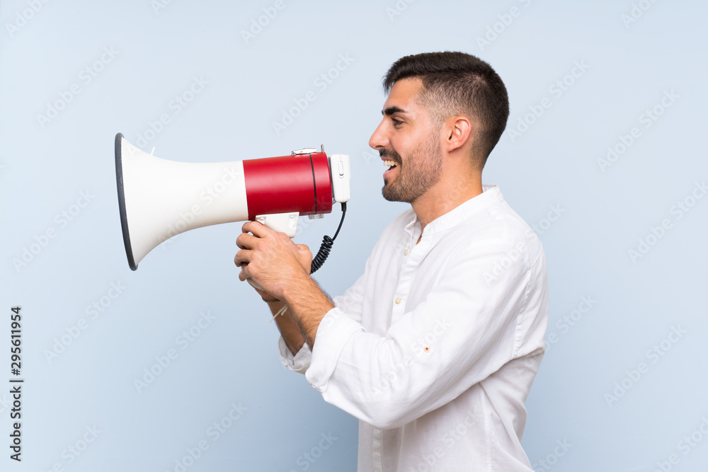 Handsome man with beard over isolated blue background shouting through a megaphone Handsome man with beard over isolated blue background shouting through a megaphone