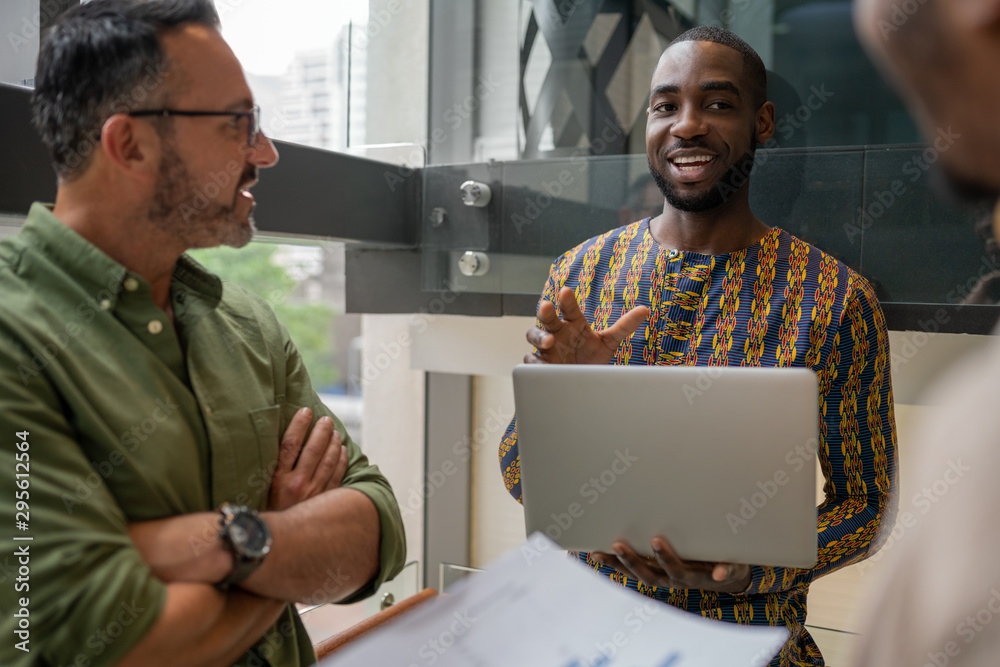 Fototapeta premium Handsome black african businessman holding laptop smiling talking to coworkers wearing African inspired pattern shirt