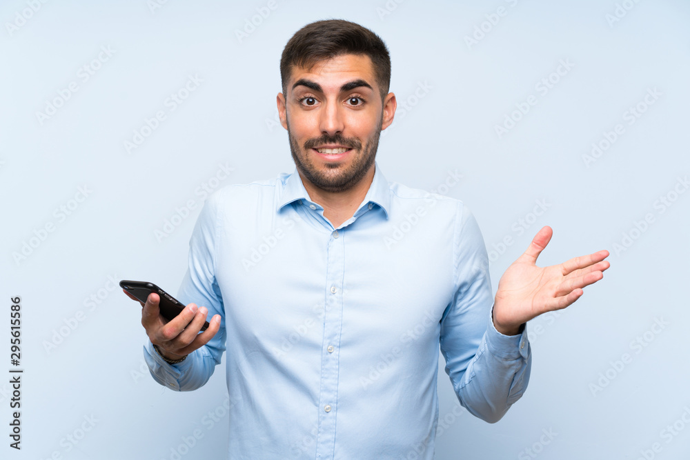 Young handsome man with his mobile over isolated blue wall with shocked facial expression Young handsome man with his mobile over isolated blue wall with shocked facial expression