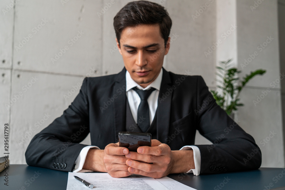 Serious attractive young businessman in dark suit holding smartphone while sitting at loft office table. Concept of internet and technology Serious attractive young businessman in dark suit holding smartphone while sitting at loft office table. Concept of internet and technology