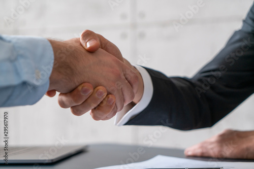 Close up of two unrecognizable businessmen shaking hands in blurred office with laptop and document. Concept of partnership and employment