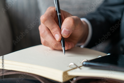 Close up of businessman hand taking notes in copybook with smartphone near it and blurred armchair in background. Concept of business lifestyle